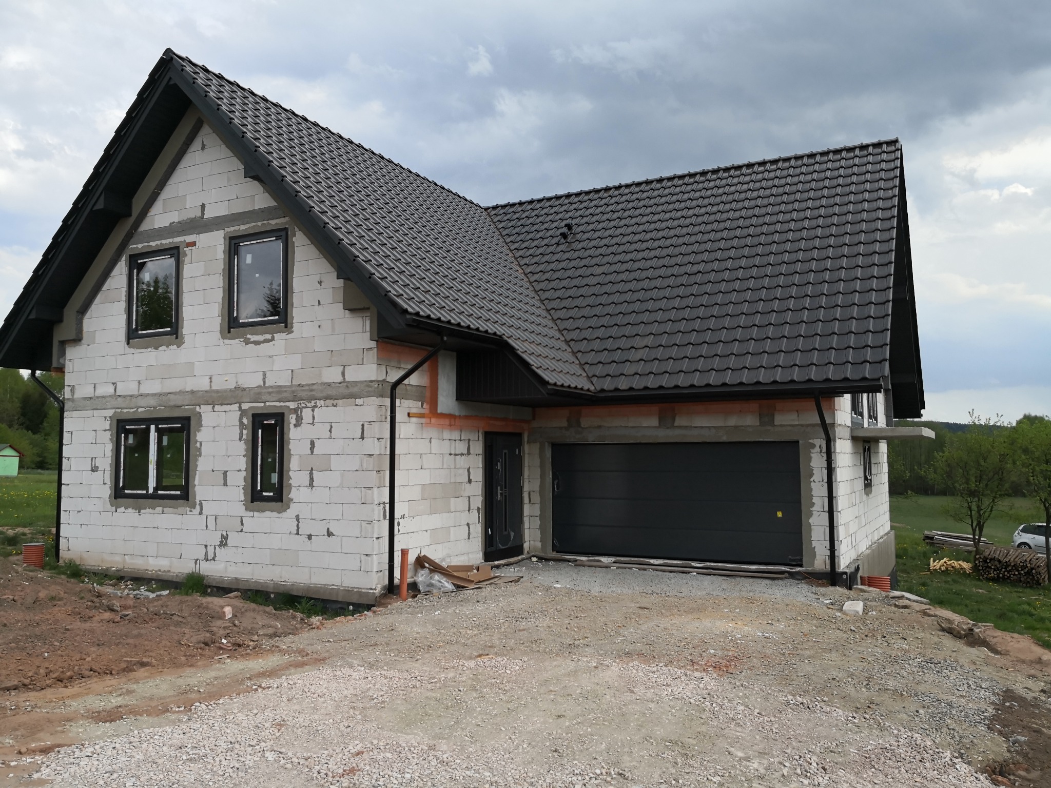 Surowy stan domu jednorodzinnego w Brynicy z zamontowanymi oknami i bramą garażową, ściany z białego betonu komórkowego, czarna dachówka i orynnowanie, teren wokół domu w trakcie prac ziemnych.