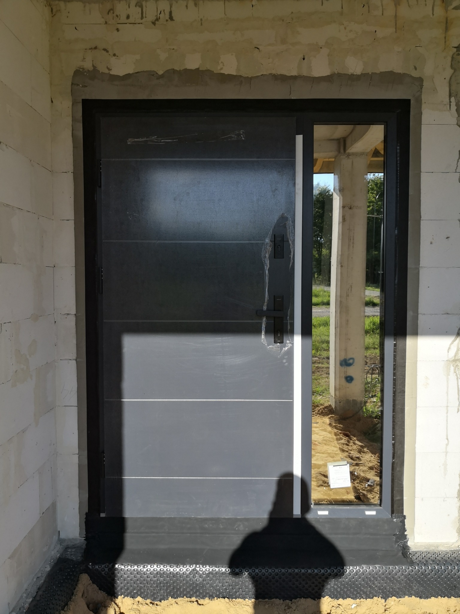 Nowe, szare drzwi wejściowe z czarną ościeżnicą, częściowo osłonięte folią, zamontowane w surowym murze z betonu komórkowego, widoczny fragment izolacji fundamentu i cień osoby robiącej zdjęcie.