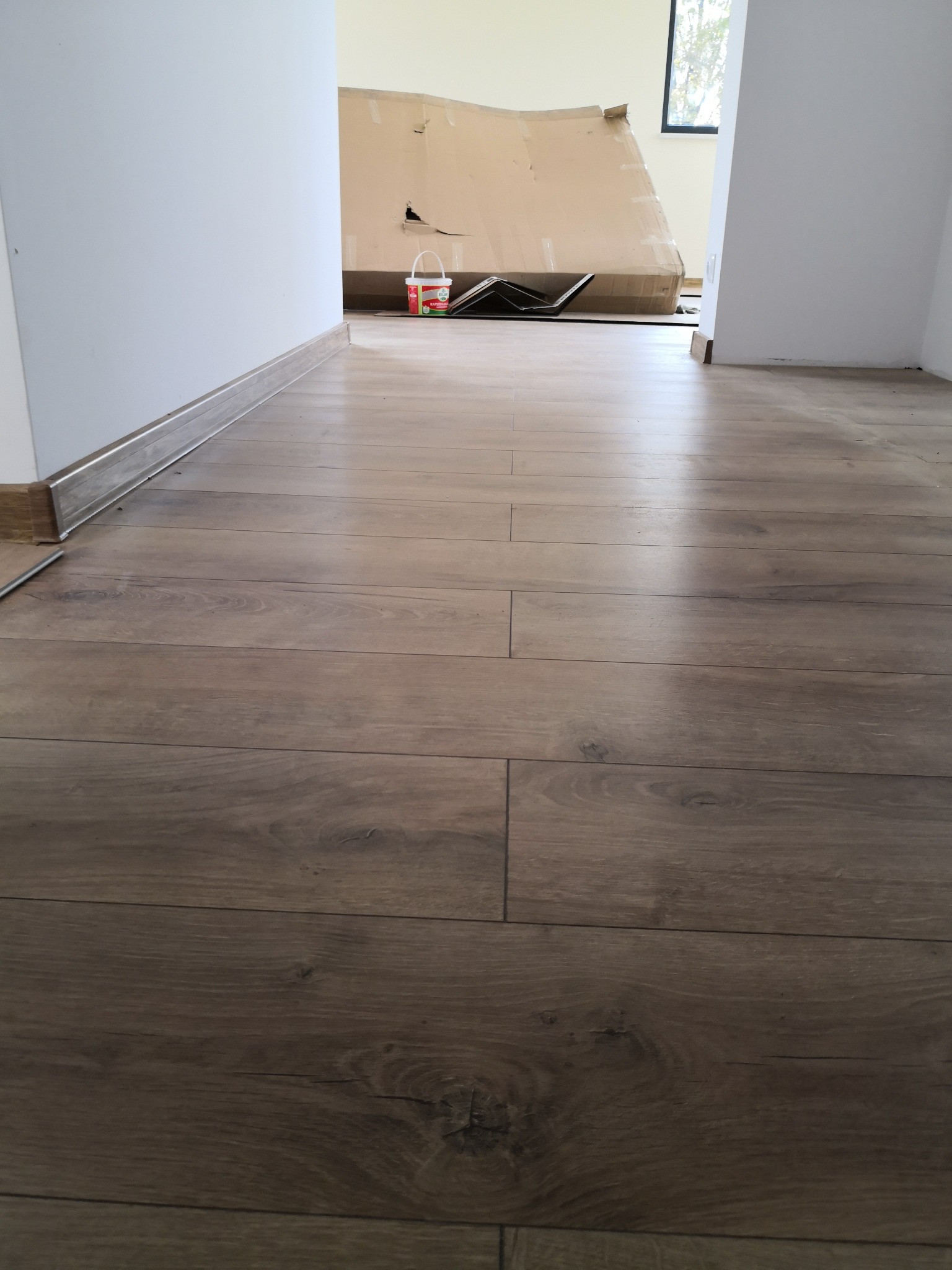 Ułożone panele podłogowe w jasnym pomieszczeniu, widoczne listwy przypodłogowe, karton i wiaderko z farbą w tle.