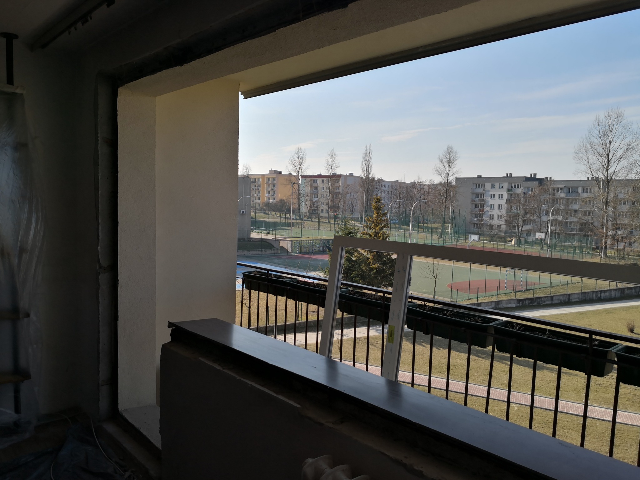 Przygotowanie do montażu okna balkonowego; widok na osiedle z boiskami sportowymi w tle, rama okienna oparta o balustradę.
