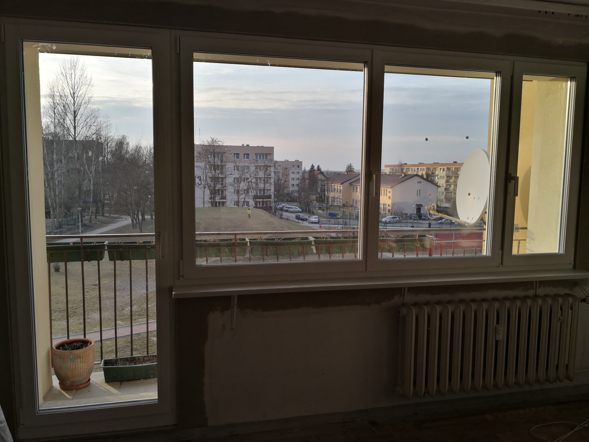 Widok z okna balkonowego na osiedle mieszkaniowe; widoczne nowe okna PCV, surowa ściana pod oknem z grzejnikiem.