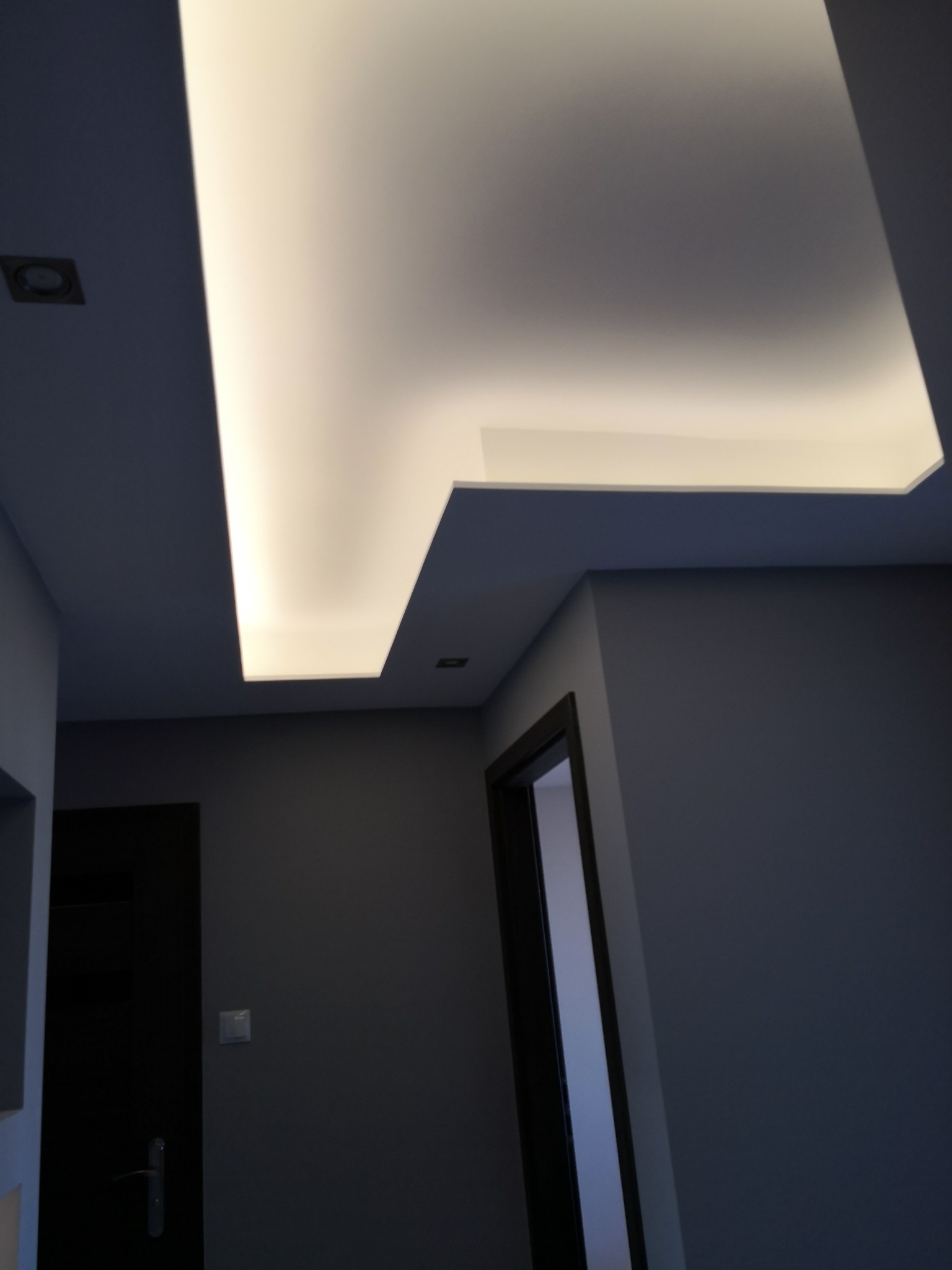 Nowoczesny sufit podwieszany z oświetleniem LED w kształcie litery L, szare ściany, widoczny fragment czarnych drzwi w pomieszczeniu.