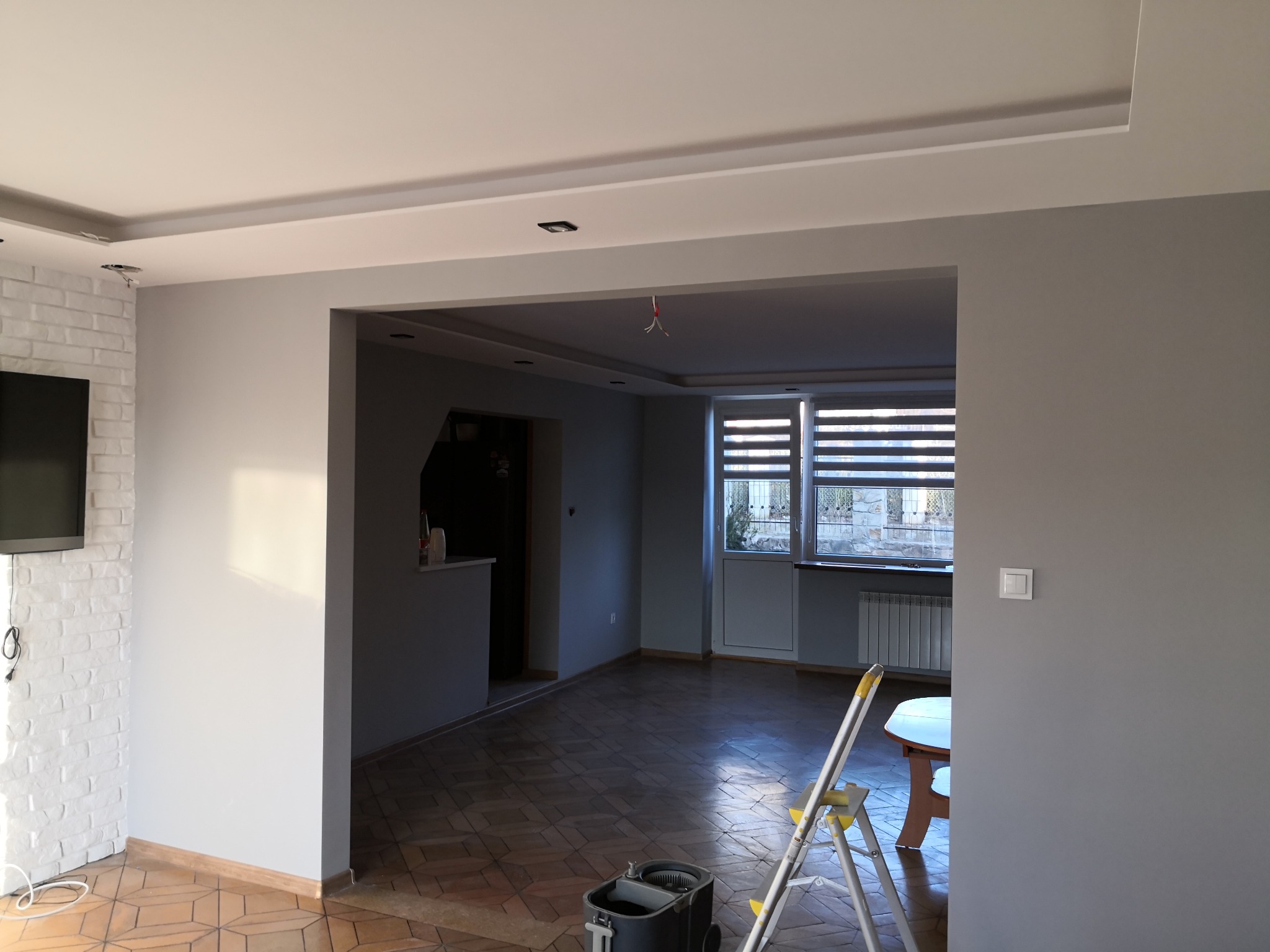 Pokój po odświeżeniu ścian szarą farbą, widoczna drabina malarska, parkiet w jodełkę, biała cegła na ścianie oraz sufit podwieszany z oświetleniem.