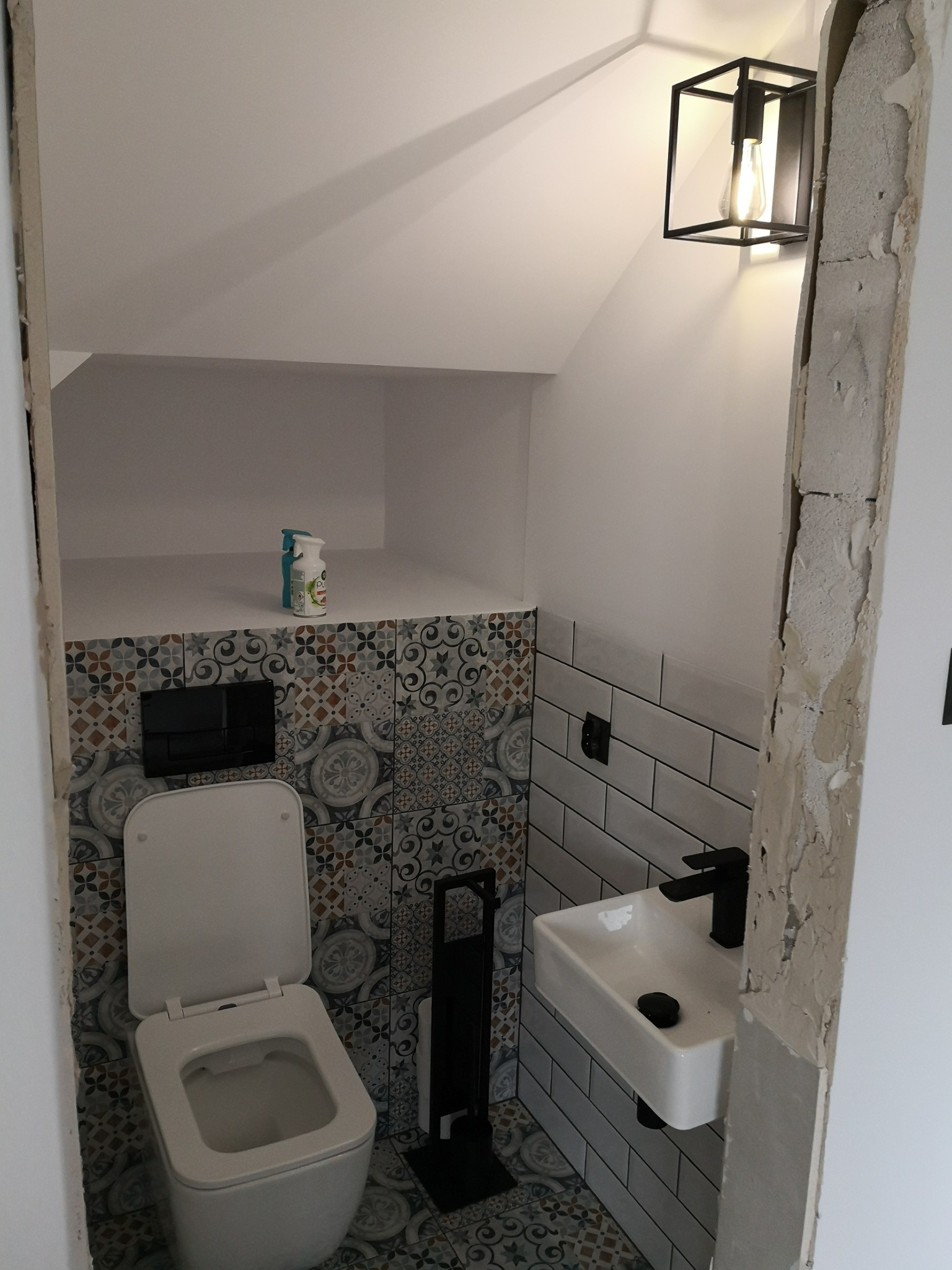 Mała łazienka na poddaszu z toaletą, umywalką i dekoracyjnymi płytkami, oświetlona nowoczesną lampą ścienną, widoczne ślady po remoncie przy ościeżnicy.