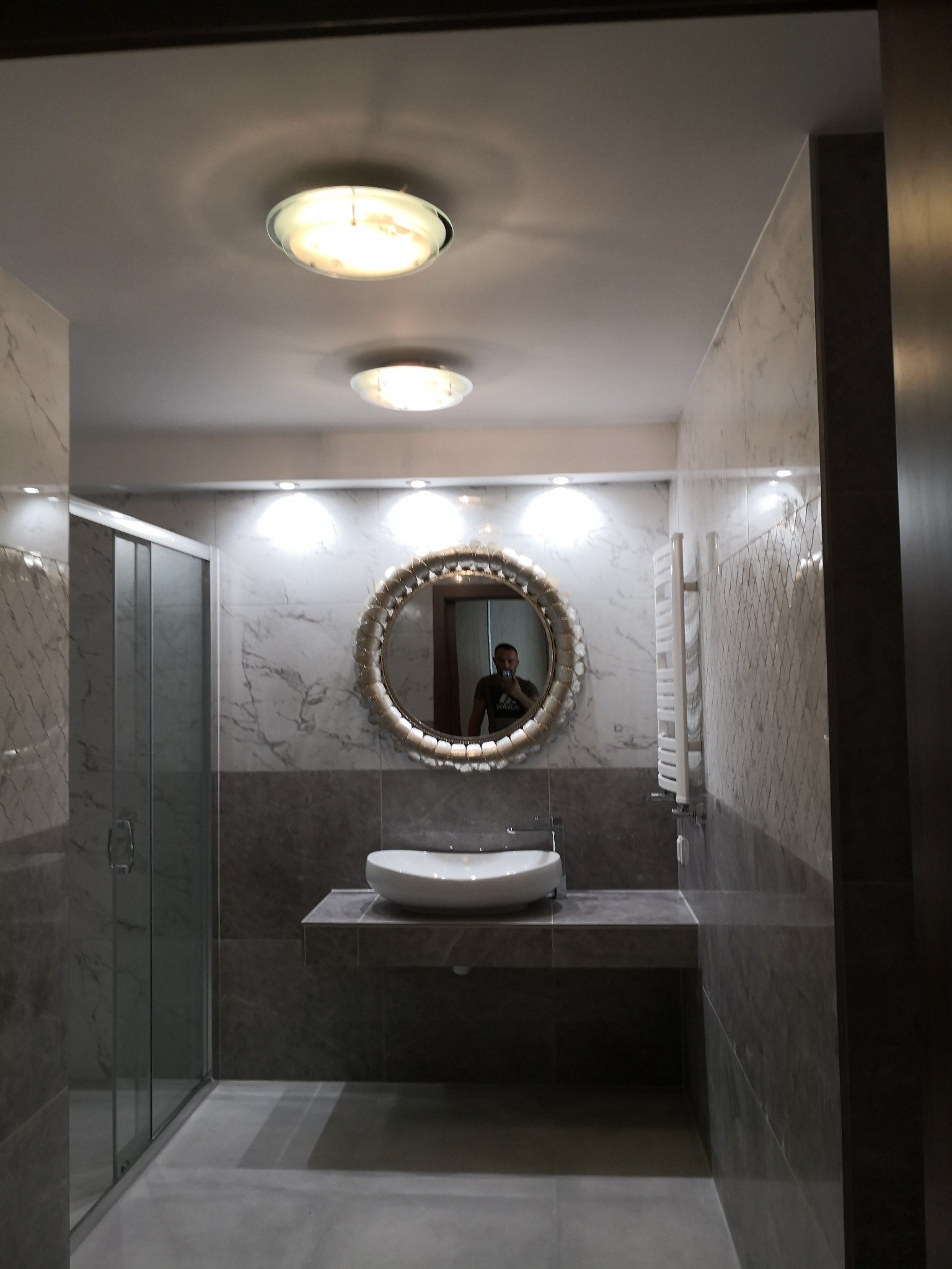 Nowoczesna łazienka w Brynicy z kabiną prysznicową, umywalką nablatową i okrągłym lustrem w ozdobnej ramie, wykończona płytkami imitującymi marmur.