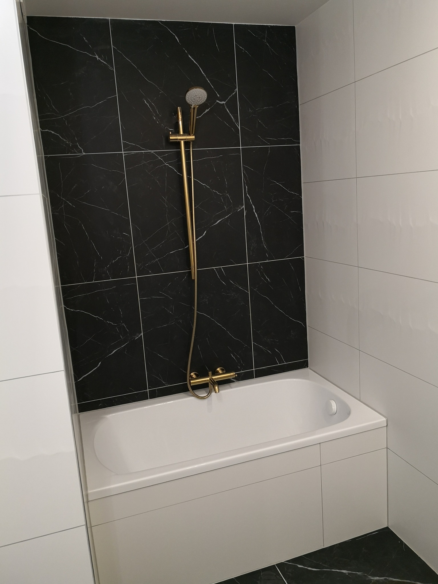 Nowoczesna łazienka z wanną obudowaną białymi płytkami, czarne marmurowe płytki na ścianie za wanną i złota armatura prysznicowa.