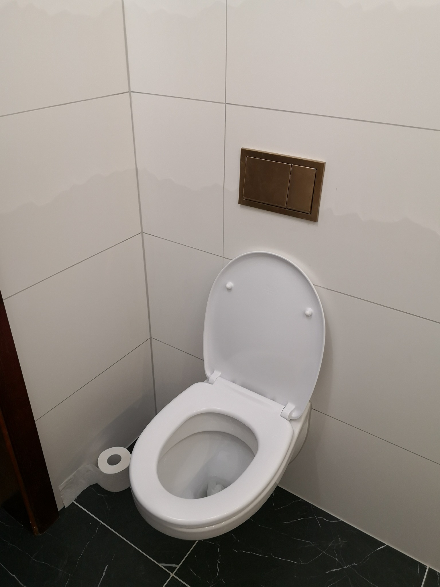 Nowoczesna toaleta z otwartą klapą, białe płytki ścienne i czarna marmurowa podłoga, obok rolka papieru toaletowego, widoczny fragment futryny drzwi.