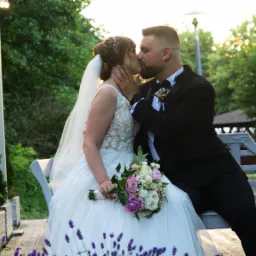 Pocałunek nowożeńców siedzących na białej ławce w plenerze, panna młoda w koronkowej sukni trzyma bukiet różowych i białych kwiatów, w tle zielone drzewa i białe kolumny.