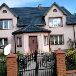 Dwupiętrowy dom z ciemną dachówką i oknami z kwiatami, ogrodzony kutym płotem z ceglanymi słupkami.