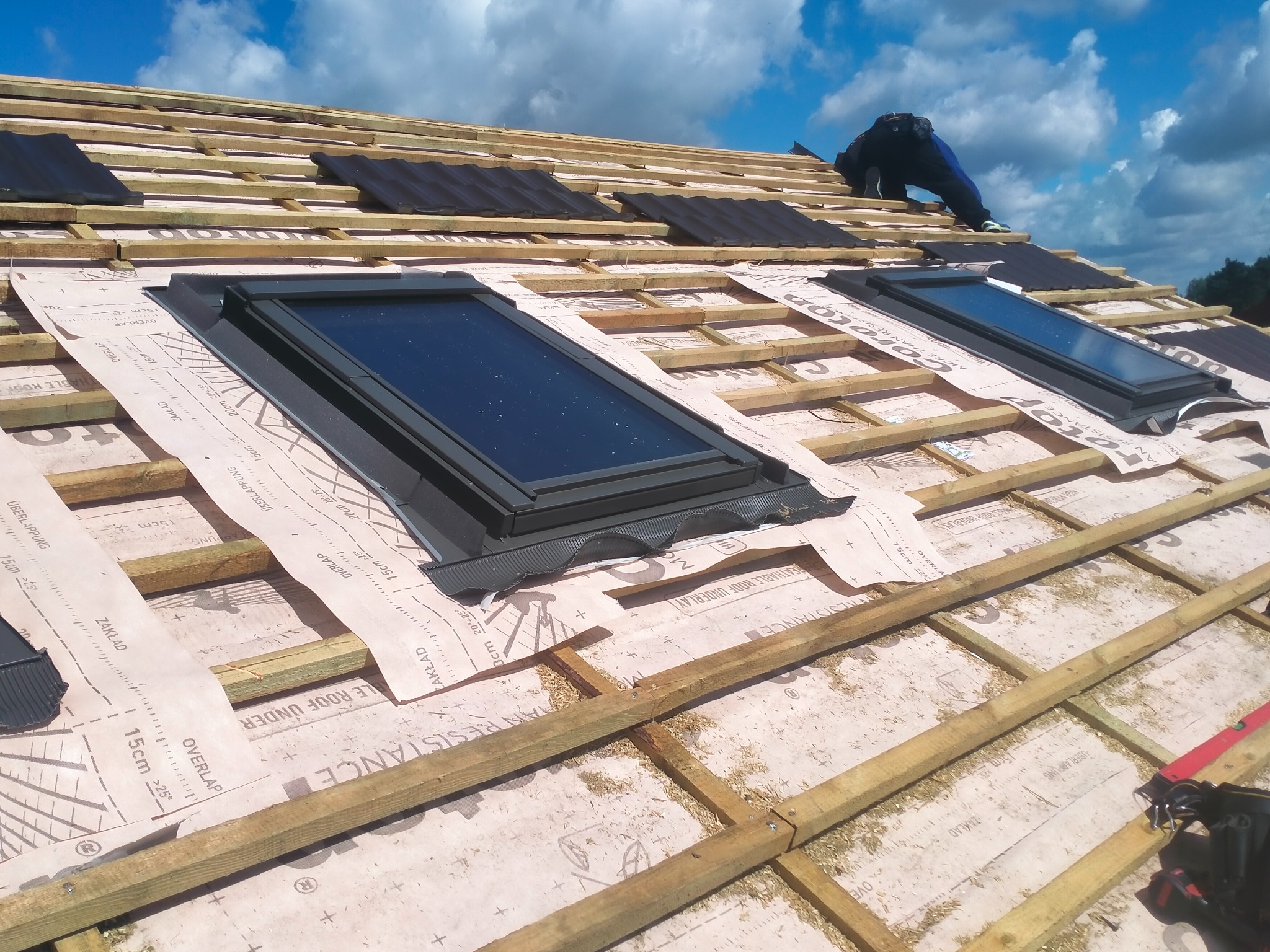 Montaż okien dachowych na konstrukcji z łat i kontrłat pokrytej membraną dachową z widocznymi oznaczeniami zakładów, częściowo pokrytej dachówką ceramiczną, z pracownikiem w tle.
