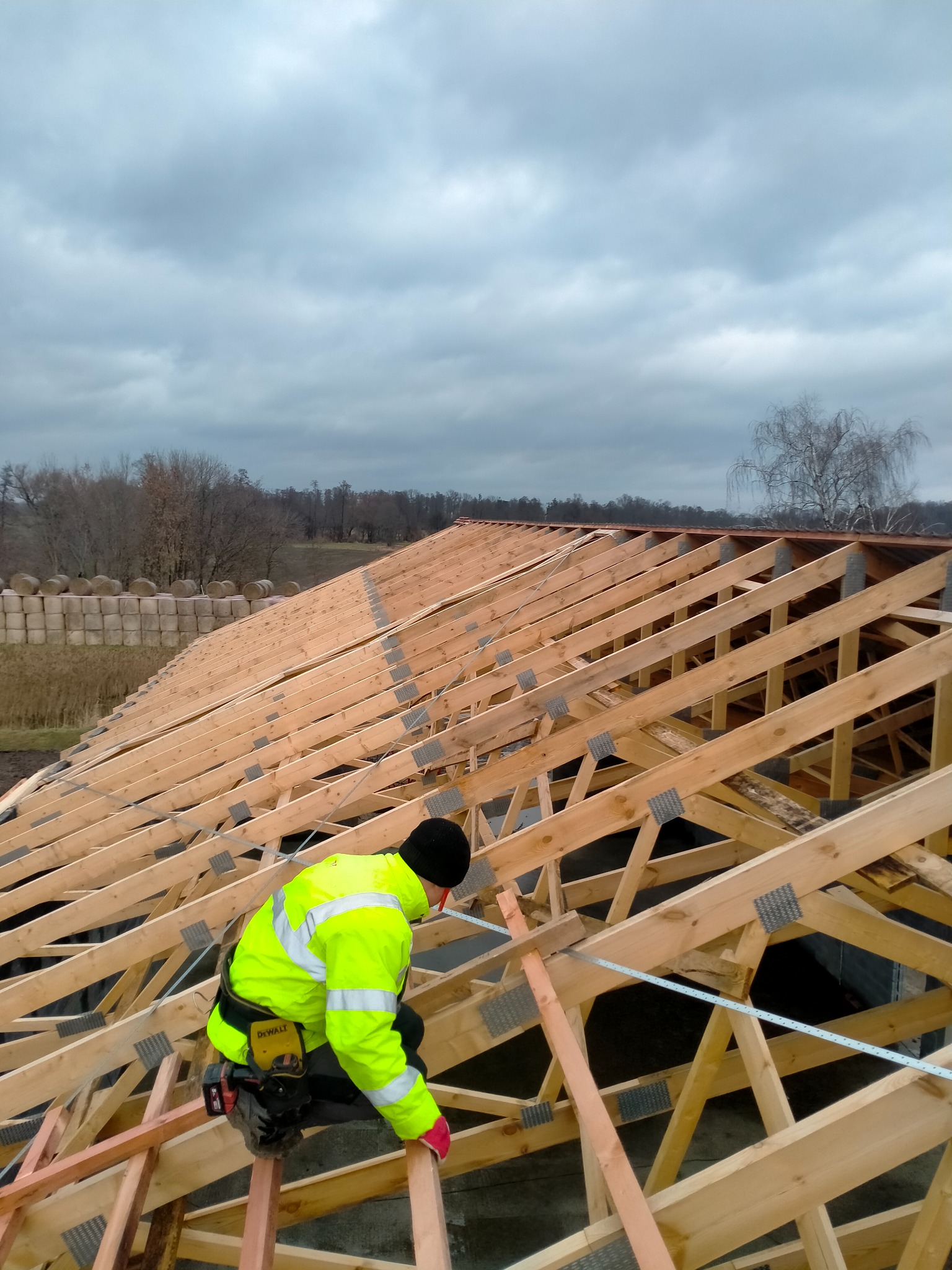 Monter w jaskrawej kurtce, klęczący na drewnianej konstrukcji, mocuje elementy więźby dachowej przy użyciu narzędzi DeWalt, w tle widoczne bele siana i pochmurne niebo.
