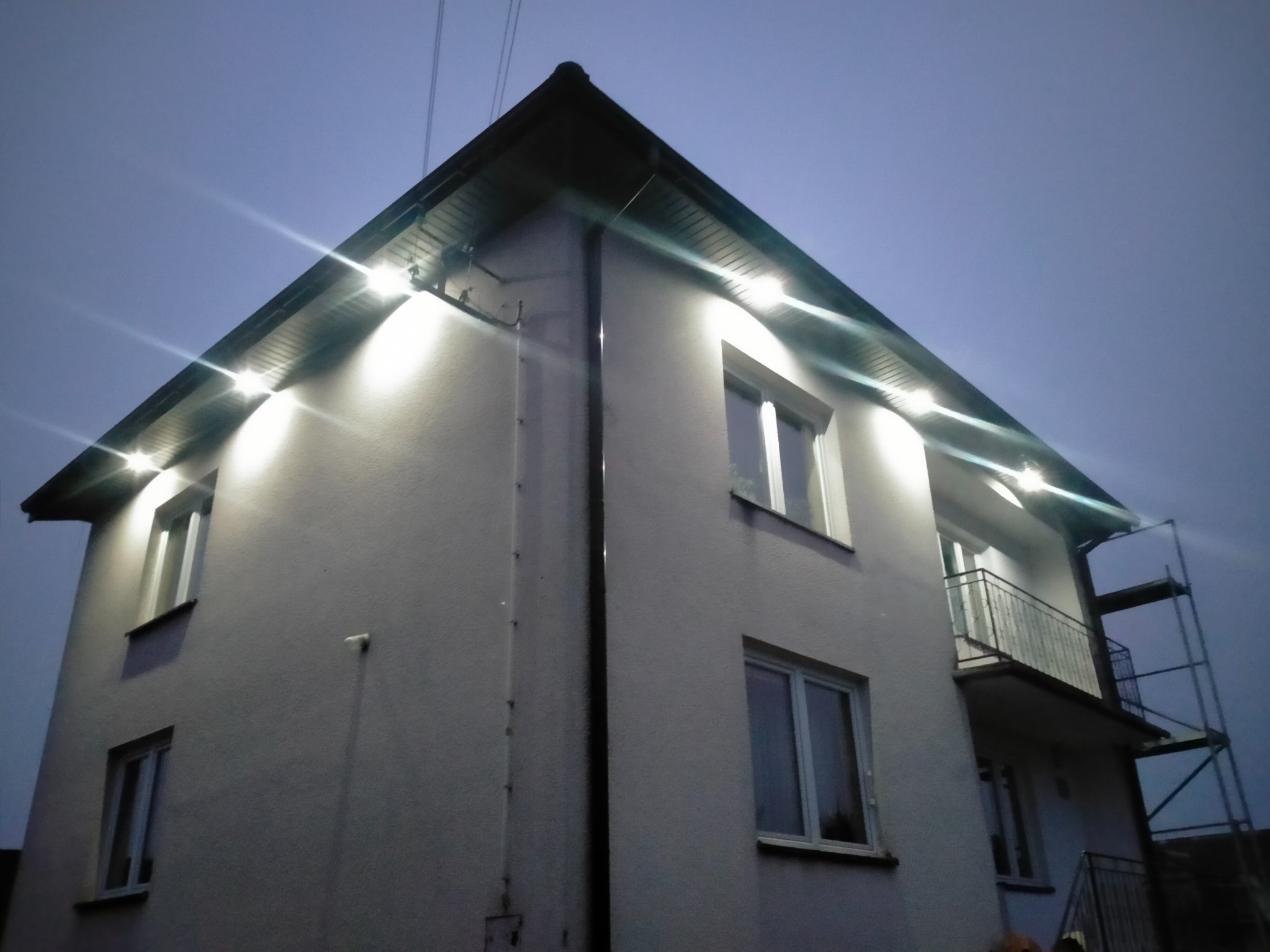 Dwupiętrowy dom z oświetleniem LED pod okapem, widoczny fragment rusztowania przy balkonie, ujęcie z dołu o zmierzchu.