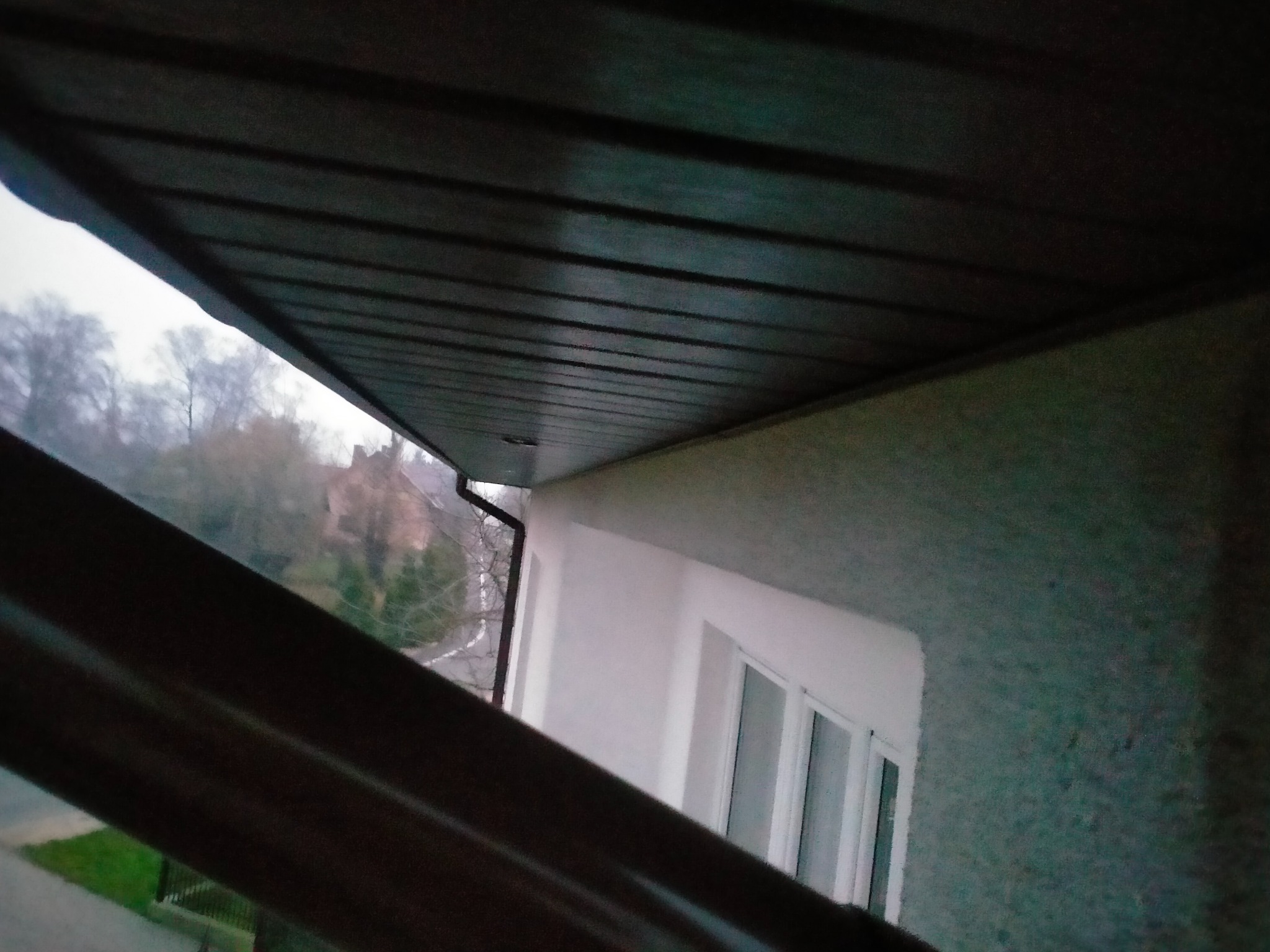 Widok z dołu na brązowe panele podbitki dachowej z zamontowaną rynną, na tle białej ściany budynku i drzew bez liści w tle.