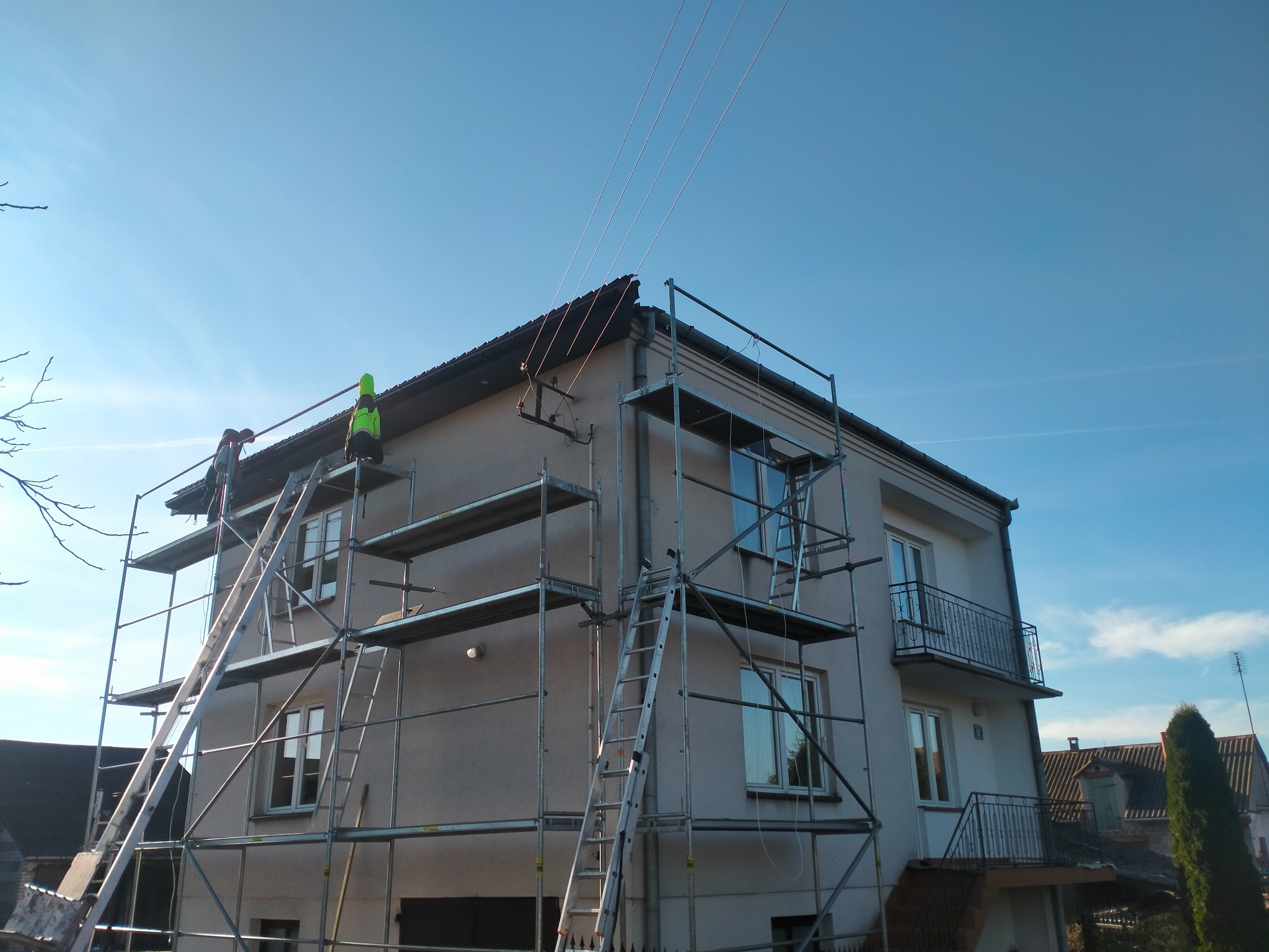 Pracownicy na rusztowaniu przy budynku dwupiętrowym, jeden w kamizelce odblaskowej, podczas prac na dachu, widoczne przewody elektryczne biegnące nad budynkiem.