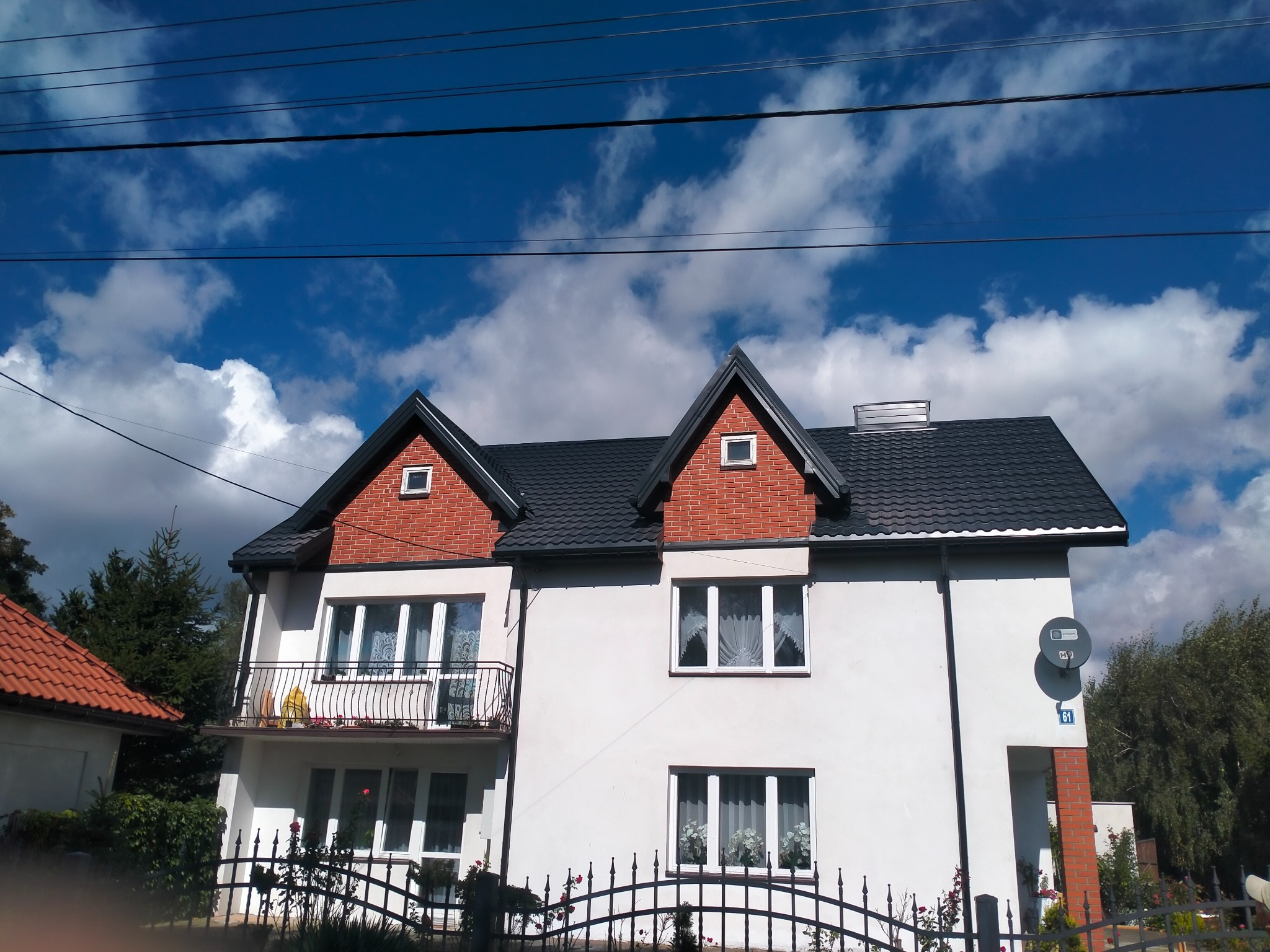 Dwupiętrowy dom z nowym, ciemnoszarym dachem z dachówki ceramicznej, widok zza metalowego ogrodzenia, błękitne niebo z chmurami w tle.