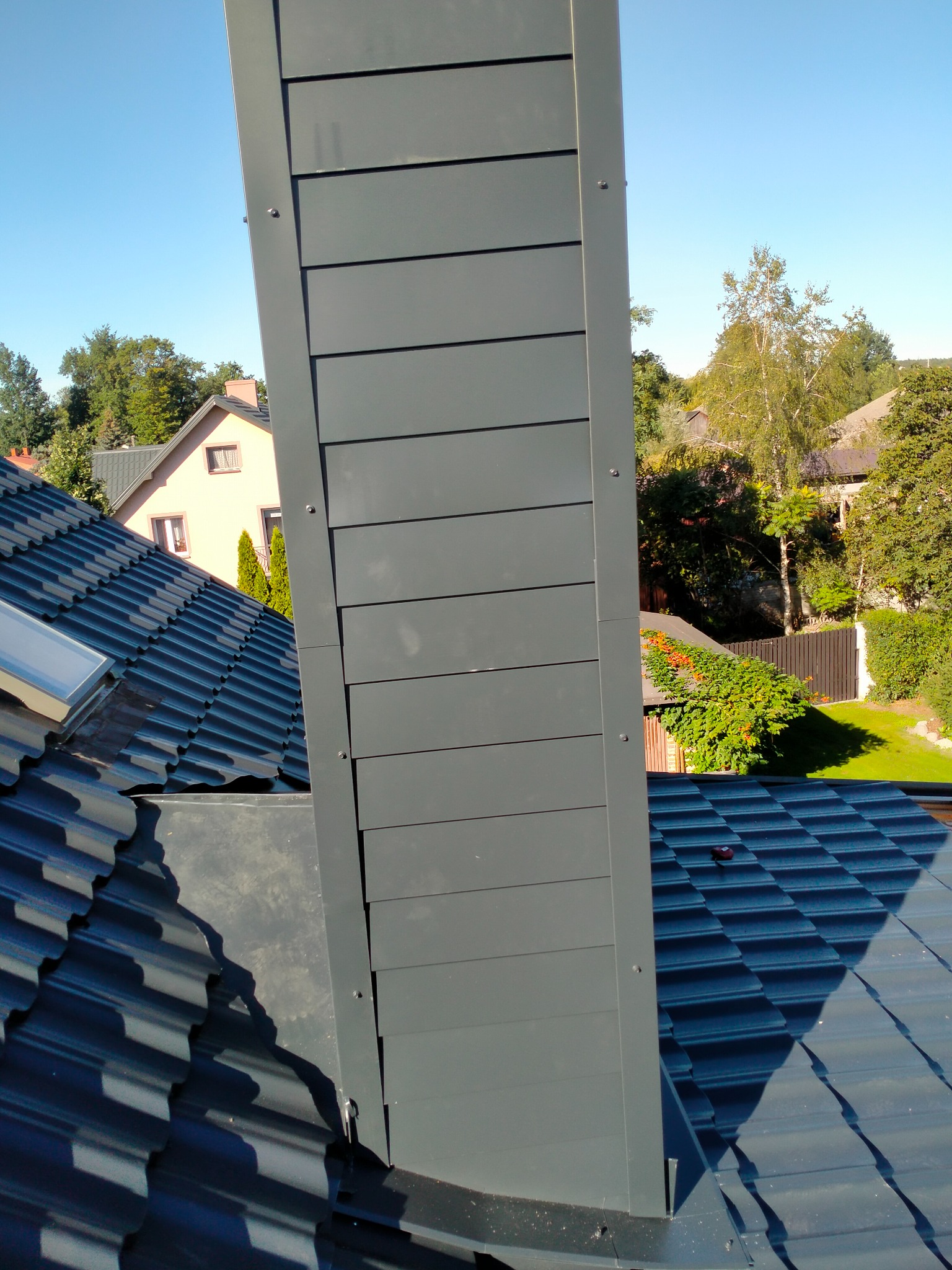 Nowoczesny komin z blachy w kolorze grafitowym, zamontowany na dachu z dachówki modułowej, widok z góry na tle zieleni drzew i zabudowań.