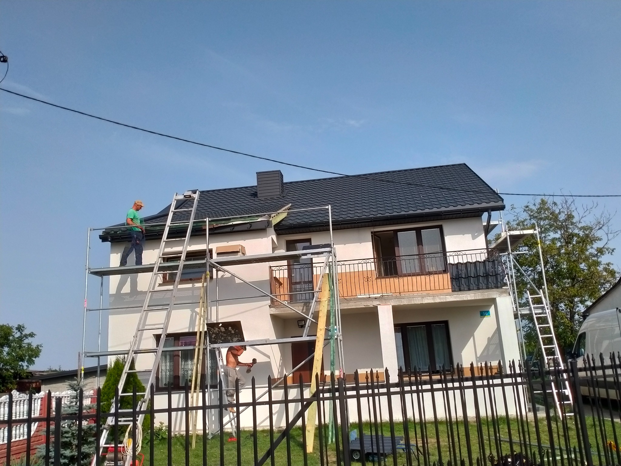 Dwóch dekarzy na rusztowaniu montuje blachodachówkę na dachu dwupiętrowego domu jednorodzinnego z czarną metalową balustradą balkonową, widok od strony ogrodu z metalowym ogrodzeniem.