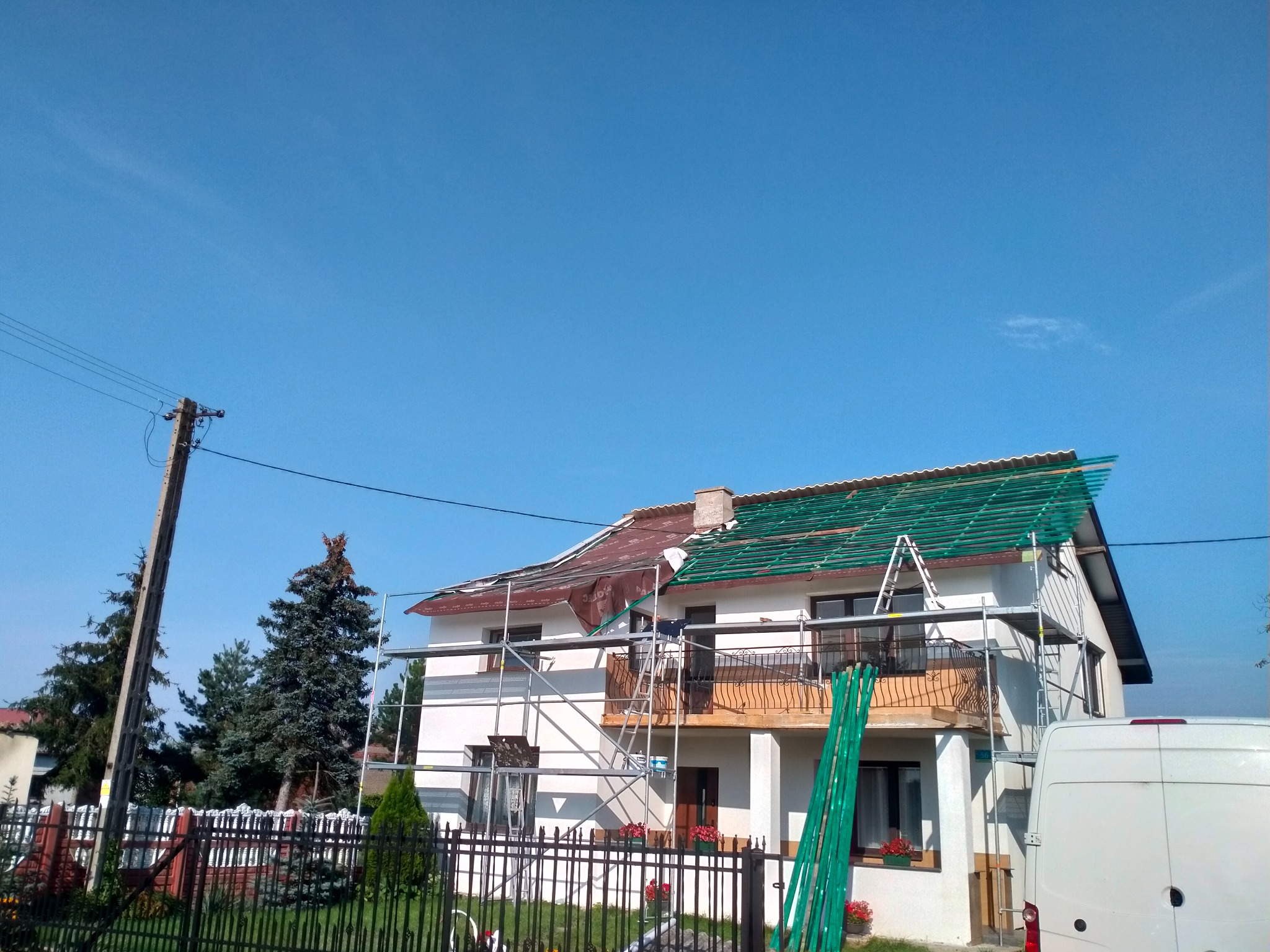 Dom jednorodzinny w trakcie prac dekarskich, widoczne rusztowania, częściowo zdjęte stare pokrycie i przygotowana konstrukcja pod nowe, błękitne niebo w tle.