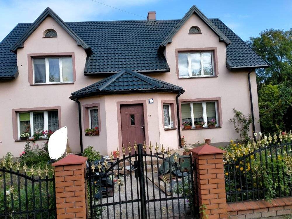 Dwupiętrowy dom z ciemną dachówką i oknami z kwiatami, ogrodzony kutym płotem z ceglanymi słupkami.