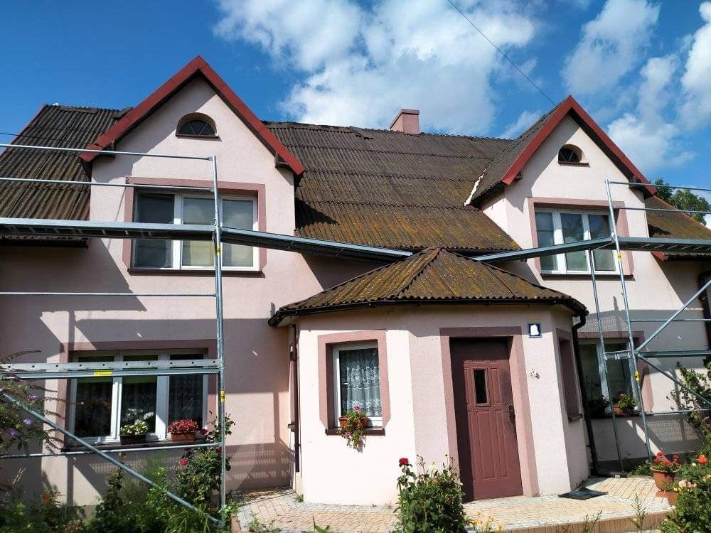 Dom z jasnoróżową elewacją i ciemnobrązowym dachem, częściowo zasłonięty rusztowaniem podczas prac remontowych.