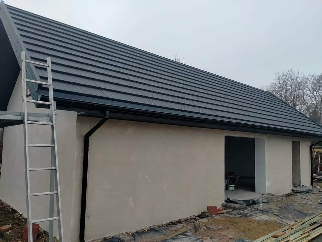 Nowy dach z czarnej dachówki ceramicznej na budynku w stanie surowym, widoczne rynny, drabina oparta o ścianę, przygotowany teren wokół budynku.