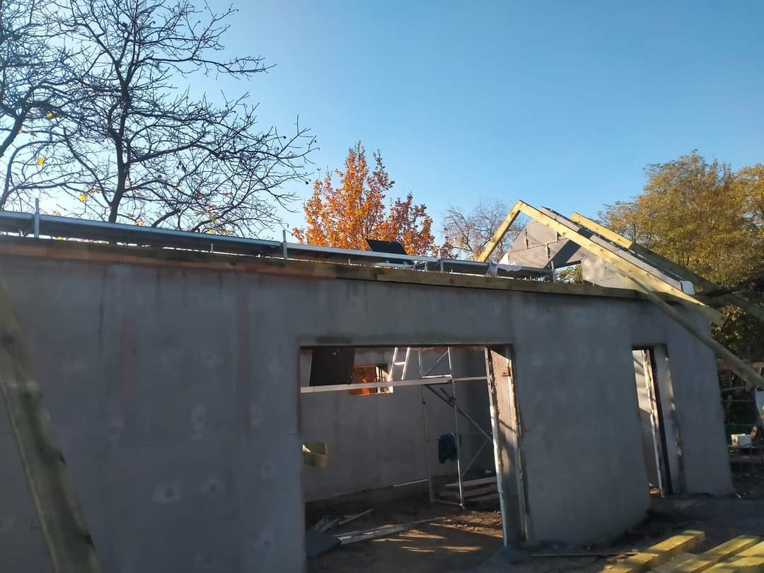 Wznoszenie więźby dachowej na surowym budynku z widocznym szarym murem i rusztowaniem wewnątrz, jesienne drzewa w tle.