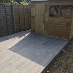 Nowoczesny taras wyłożony szarymi płytami z widoczną drewnianą altaną z oknami z abstrakcyjnymi wzorami i drewnianym ogrodzeniem w tle.