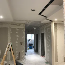 Wnętrze w trakcie prac wykończeniowych: ściany pokryte płytami gipsowo-kartonowymi z widocznymi łączeniami, sufit z nietypowym wykończeniem z płyty gipsowej, drabina i narzędzia na podłodze.