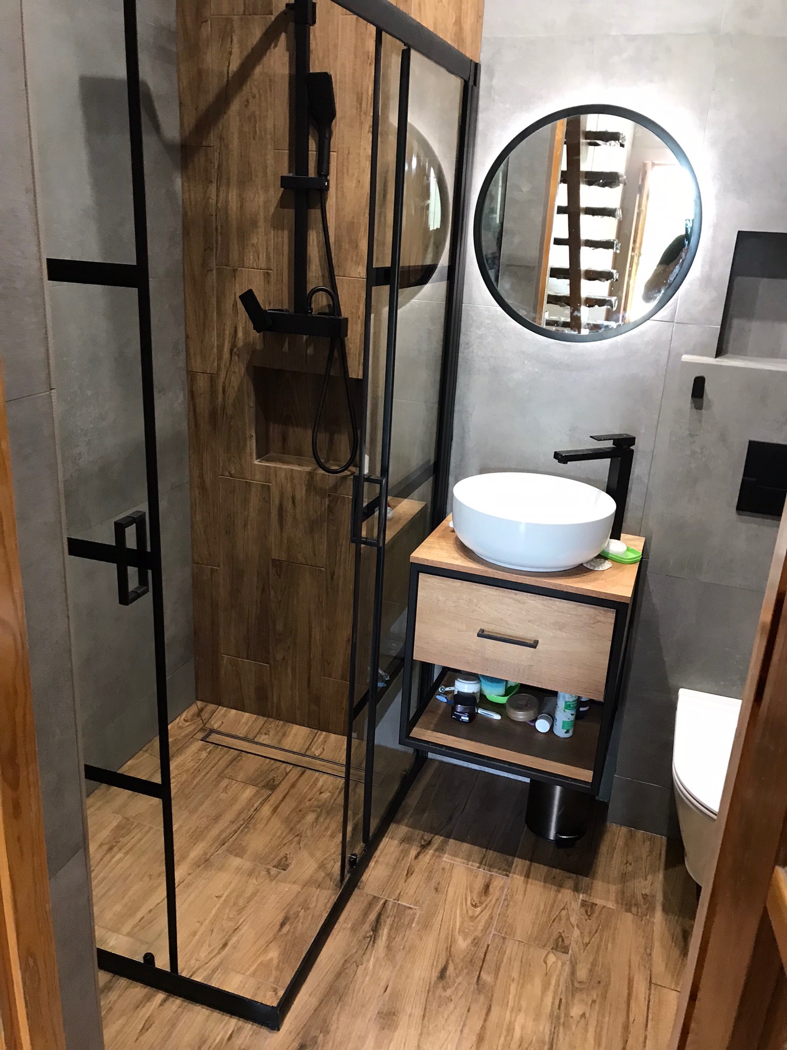 Nowoczesna łazienka z kabiną prysznicową typu walk-in z czarnymi profilami, okrągłym lustrem z podświetleniem, drewnianą szafką pod umywalkę z czarnymi elementami i drewnopodobną glazurą.