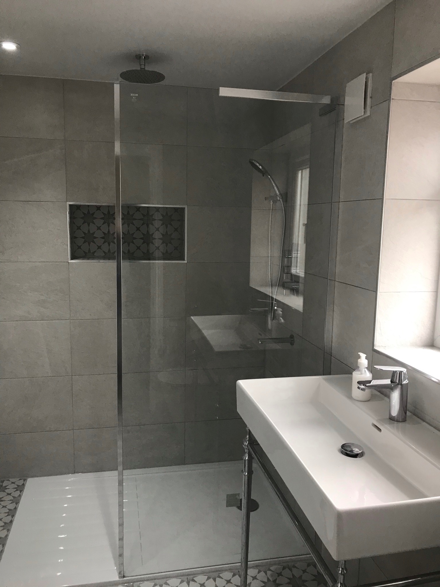 Nowoczesna łazienka z szarą płytką, kabiną prysznicową typu walk-in, umywalką na chromowanym stelażu i dekoracyjną wnęką z mozaiką.