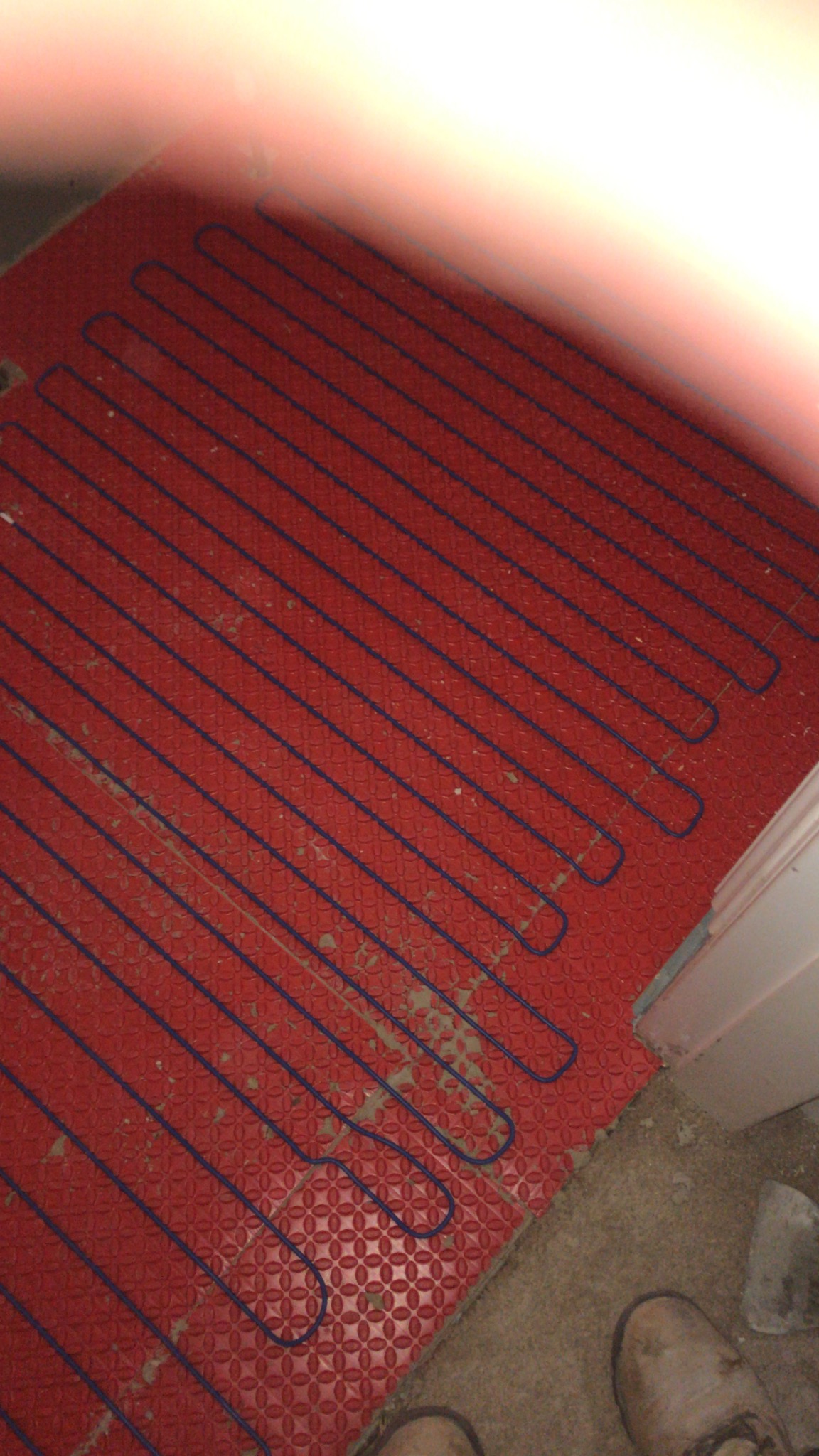 Czerwona mata grzewcza podłogowa z niebieskimi kablami ułożona na podłożu z widocznymi śladami zaprawy, częściowo przykryta butem, przed montażem wykończenia podłogi.