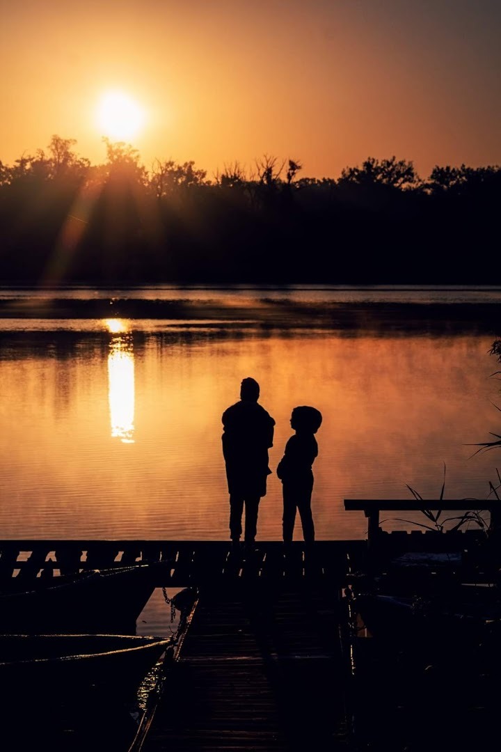 Sylwetki dorosłego i dziecka stojące na drewnianym pomoście nad jeziorem podczas wschodu słońca, z odbijającym się światłem w wodzie i zarysem lasu w tle.