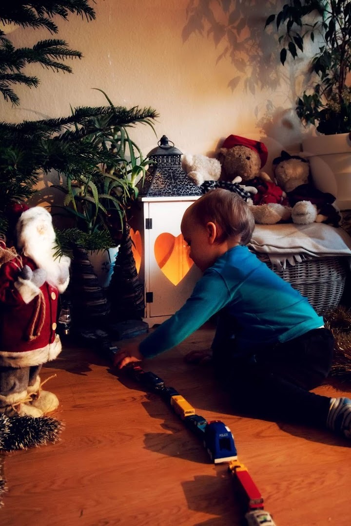 Chłopiec w niebieskiej bluzce bawi się na podłodze pociągiem z wagonów, w tle choinka, lampion z wyciętym sercem i pluszowe misie.