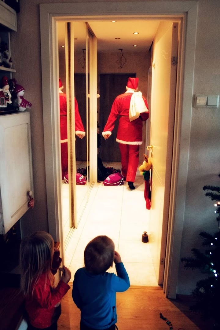 Dwoje dzieci obserwuje postać w stroju Świętego Mikołaja wchodzącą do pokoju, widoczne odbicie w lustrze, torba z prezentami i dekoracje świąteczne.