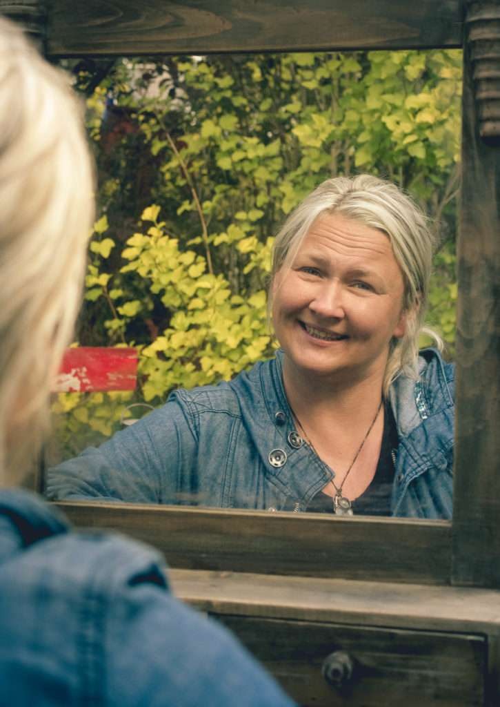 Uśmiechnięta niania w dżinsowej kurtce, widziana w lustrze na tle zielonych krzewów, z widoczną częścią odbicia jej twarzy.