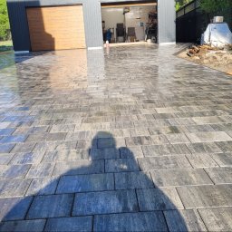 Usługi Brukarskie Piotr Całka - Świeżo ułożona, wilgotna kostka brukowa w odcieniach szarości prowadząca do garażu z drewnianymi drzwiami. Widoczny cień osoby robiącej zdjęcie i fragment schodów.