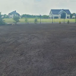 Przygotowany teren pod trawnik z widocznymi śladami po maszynach, obsadzony młodymi drzewkami iglastymi i liściastymi wzdłuż ogrodzenia panelowego na betonowej podmurówce, w tle zabudowa...