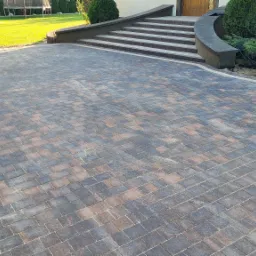 Nowa, wielobarwna nawierzchnia z kostki brukowej prowadząca do schodów wejściowych domu, z dekoracyjnym obrzeżem i zadbanym trawnikiem w tle.