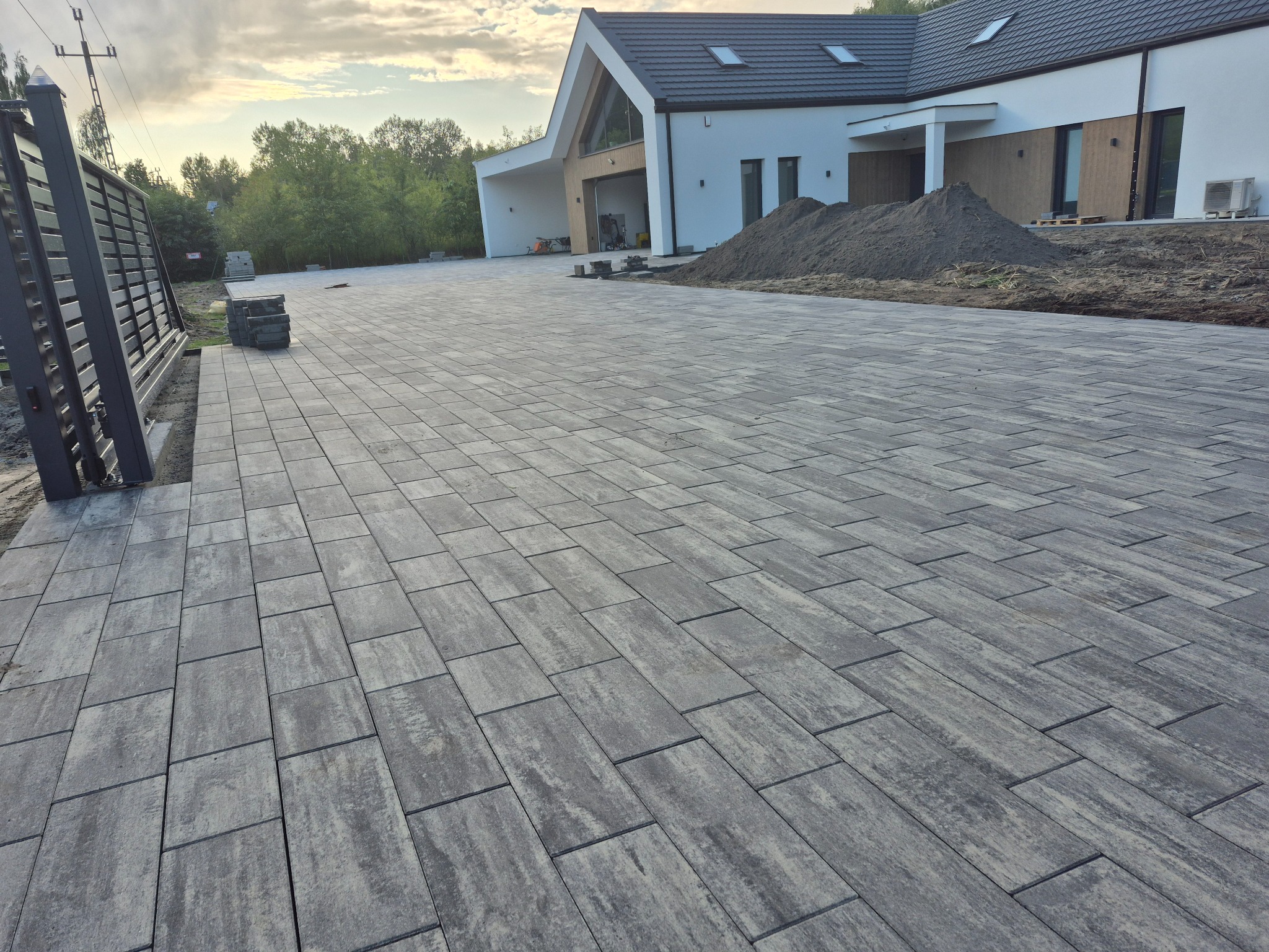 Ułożona, szara kostka brukowa prowadząca do nowoczesnego domu z białymi ścianami i ciemnym dachem. Widoczna brama wjazdowa i sterta ziemi obok domu.