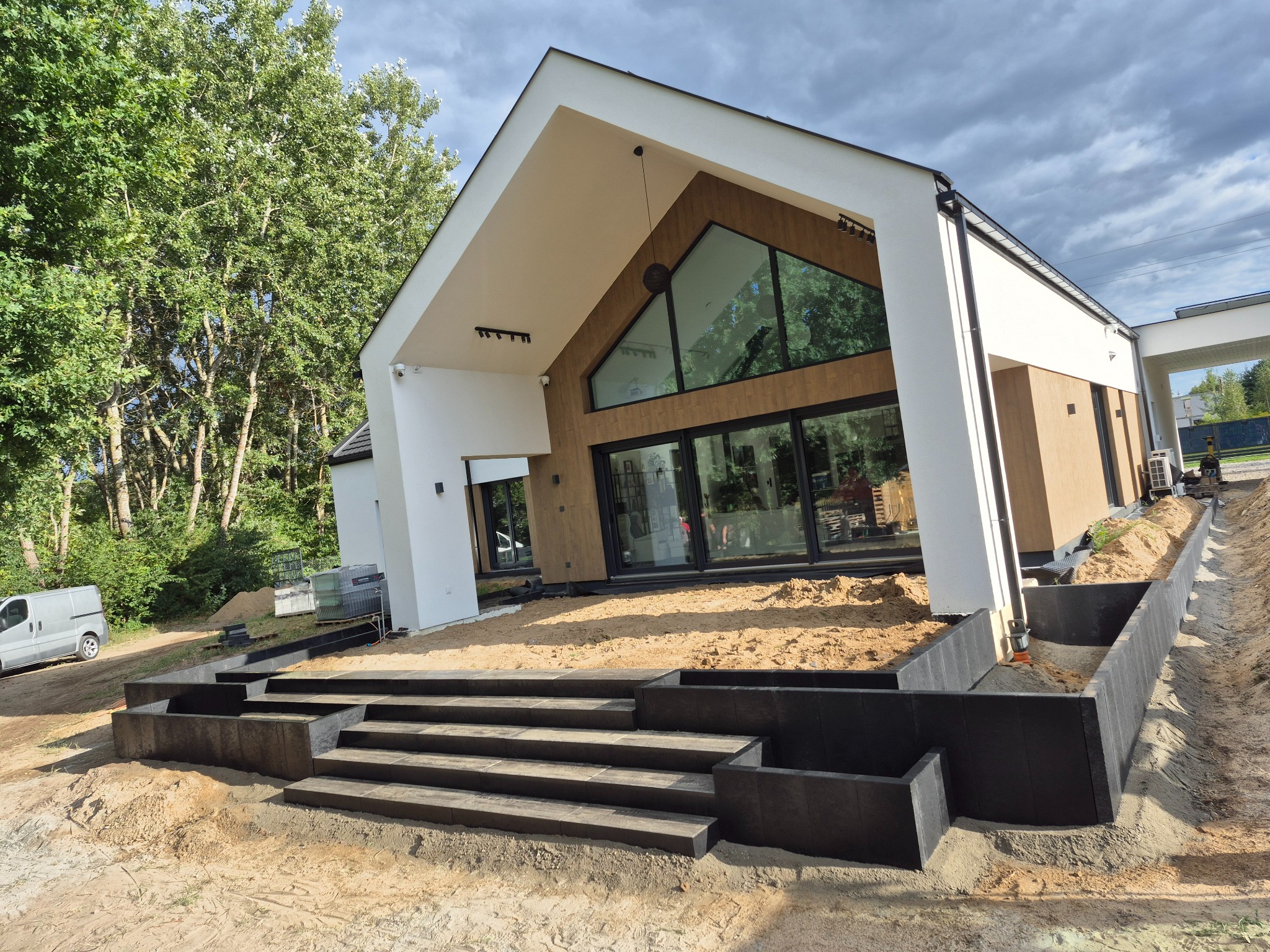 Nowoczesny dom z czarnymi schodami i obrzeżami z kostki. Widoczny teren przygotowany pod dalsze prace brukarskie, z zaparkowanym vanem w tle.