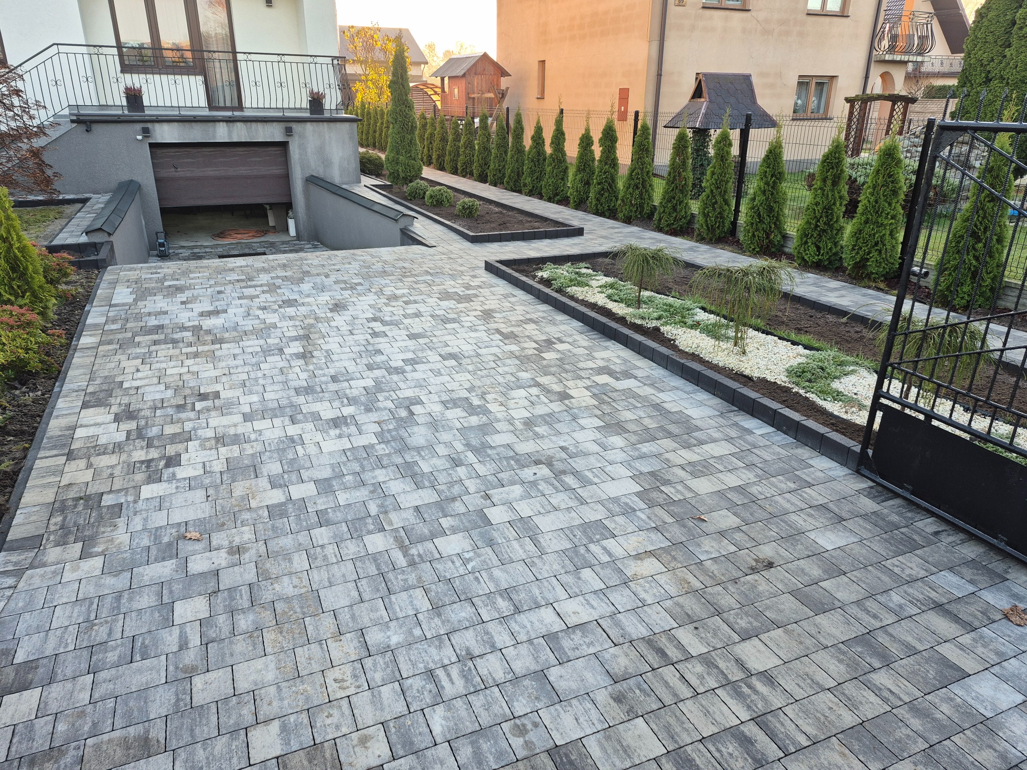 Ułożona szara kostka brukowa na podjeździe do garażu, z rabatami i tujami w tle. Widok na dom z balkonem i ogrodzenie.
