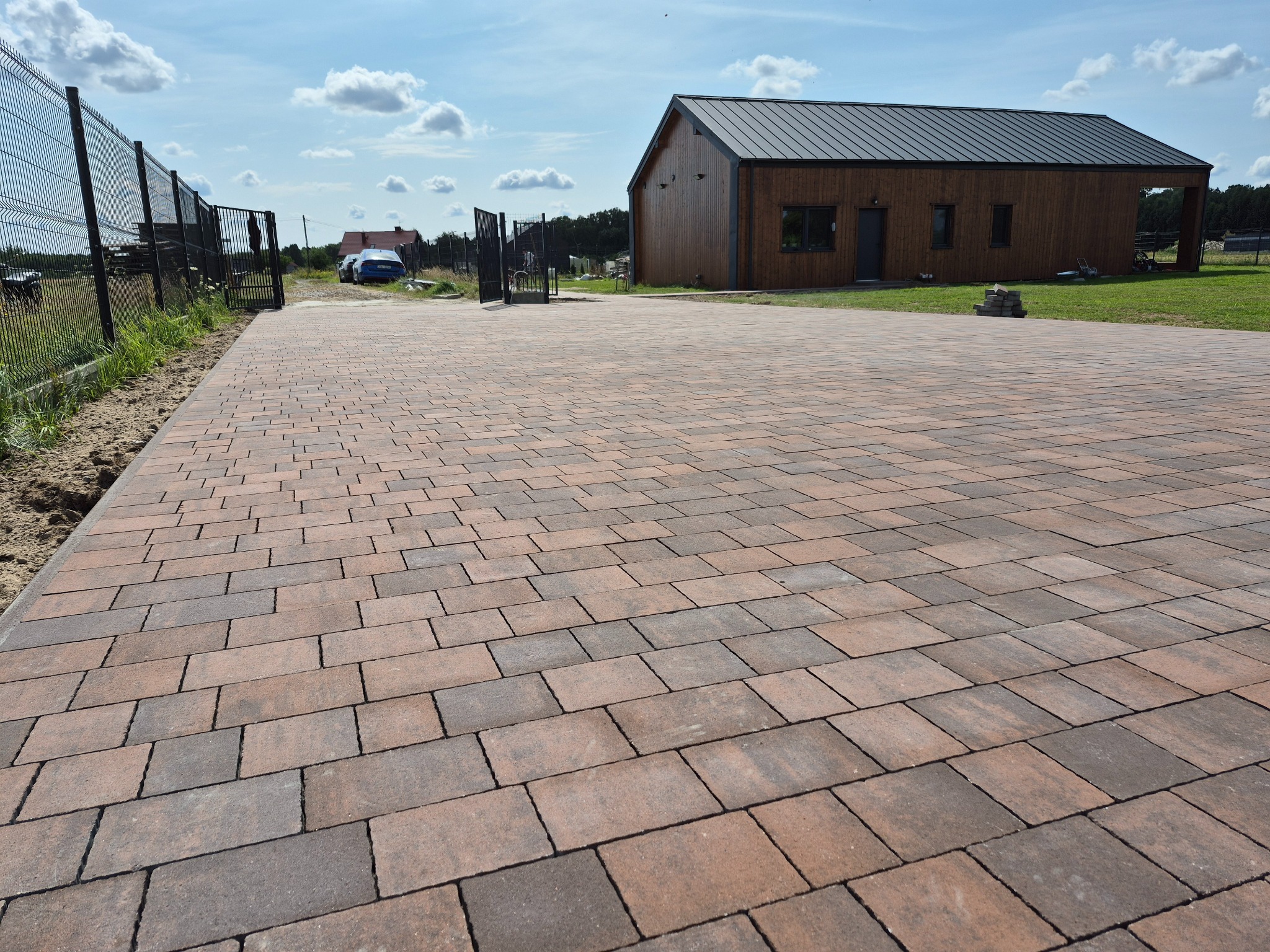 Ułożona kostka brukowa w odcieniach brązu i szarości prowadząca do nowoczesnego domu z drewnianą elewacją i metalowym dachem, widok z perspektywy.