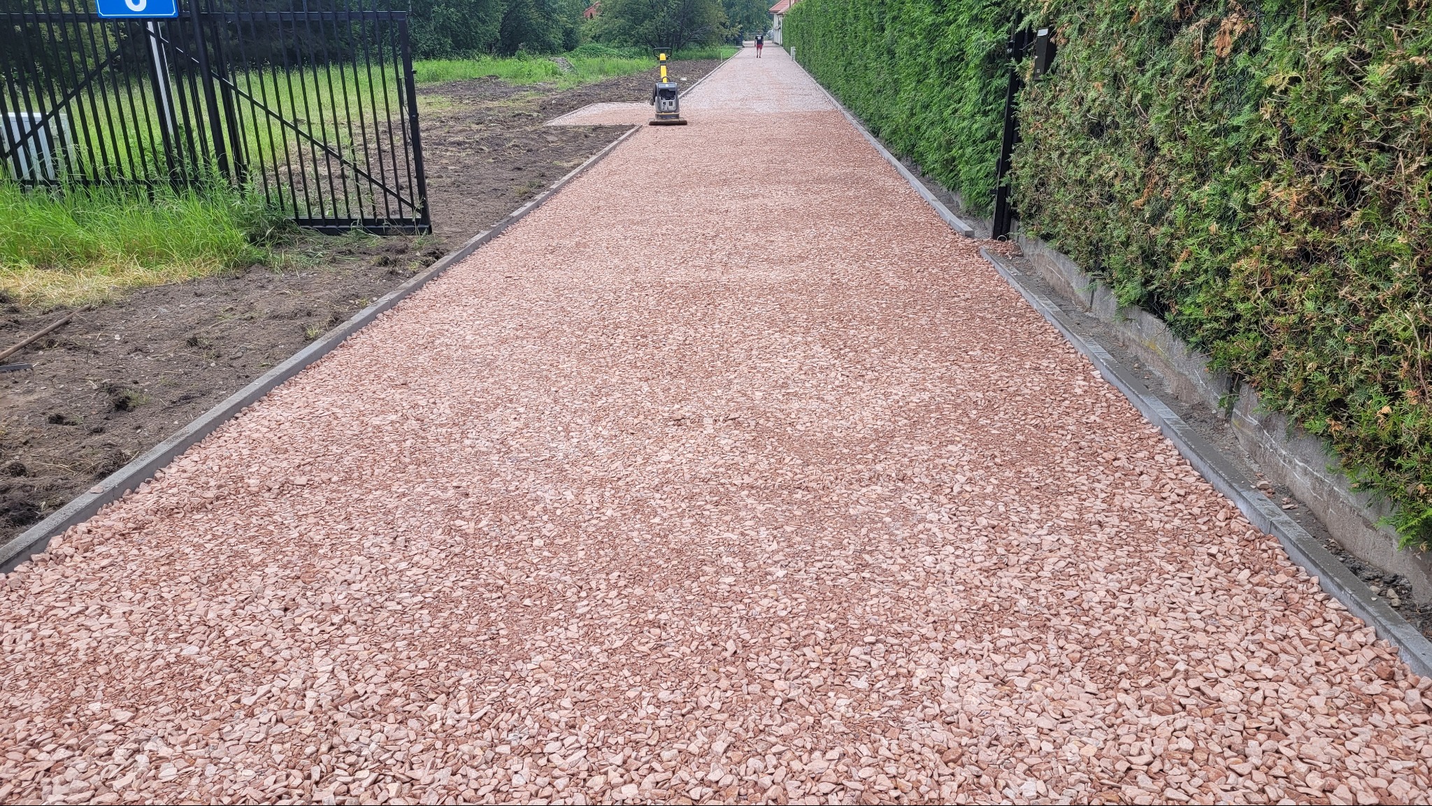 Utwardzona ścieżka z różowego kruszywa, obrzeża z szarej kostki, w tle maszyna do ubijania i osoba w oddali. Po prawej żywopłot, po lewej brama.