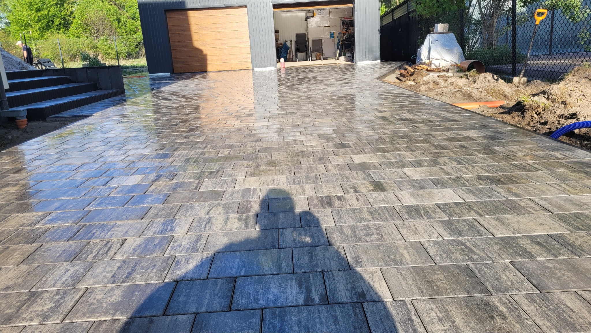 Świeżo ułożona, wilgotna kostka brukowa w odcieniach szarości prowadząca do garażu z drewnianymi drzwiami. Widoczny cień osoby robiącej zdjęcie i fragment schodów.