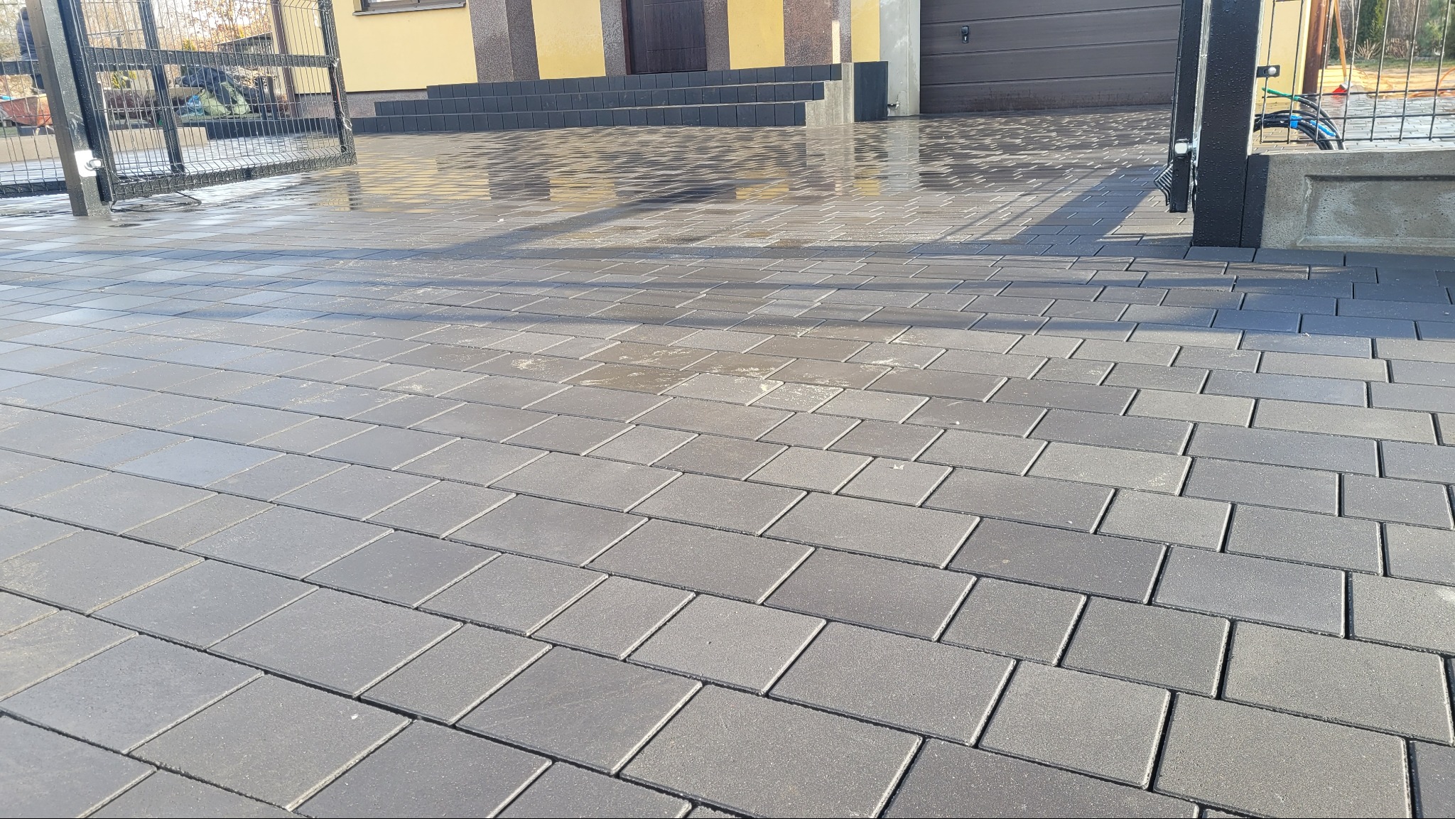 Szara kostka brukowa ułożona na podjeździe, częściowo mokra, z widoczną bramą w tle i fragmentem elewacji budynku. Regularny wzór ułożenia kostki.