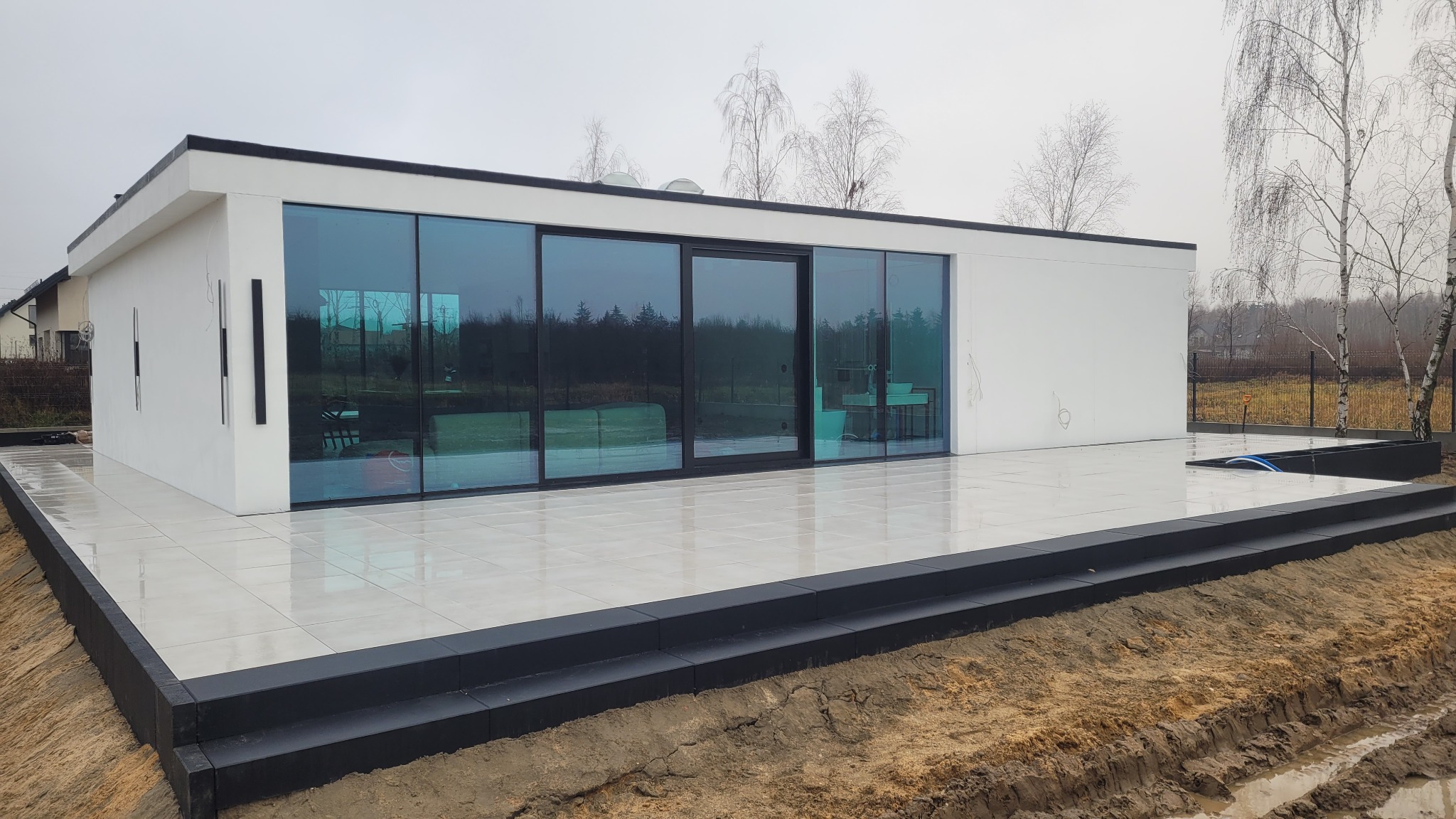 Nowoczesny, parterowy budynek z dużymi, przesuwnymi oknami i tarasem wyłożonym jasnymi płytkami. Widoczne obrzeża tarasu z ciemnej kostki i przygotowany teren wokół.