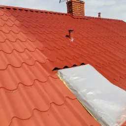 Czerwony dach z blachy trapezowej po malowaniu, z zabezpieczonym oknem dachowym folią i taśmą, widoczny komin z cegły i antena telewizyjna