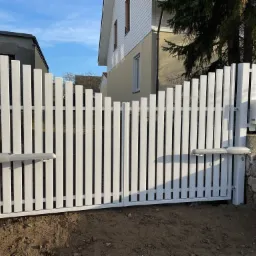 Automatyczna, biała brama wjazdowa z pionowych paneli, zamontowana na posesji z widocznym domem i murem oporowym.