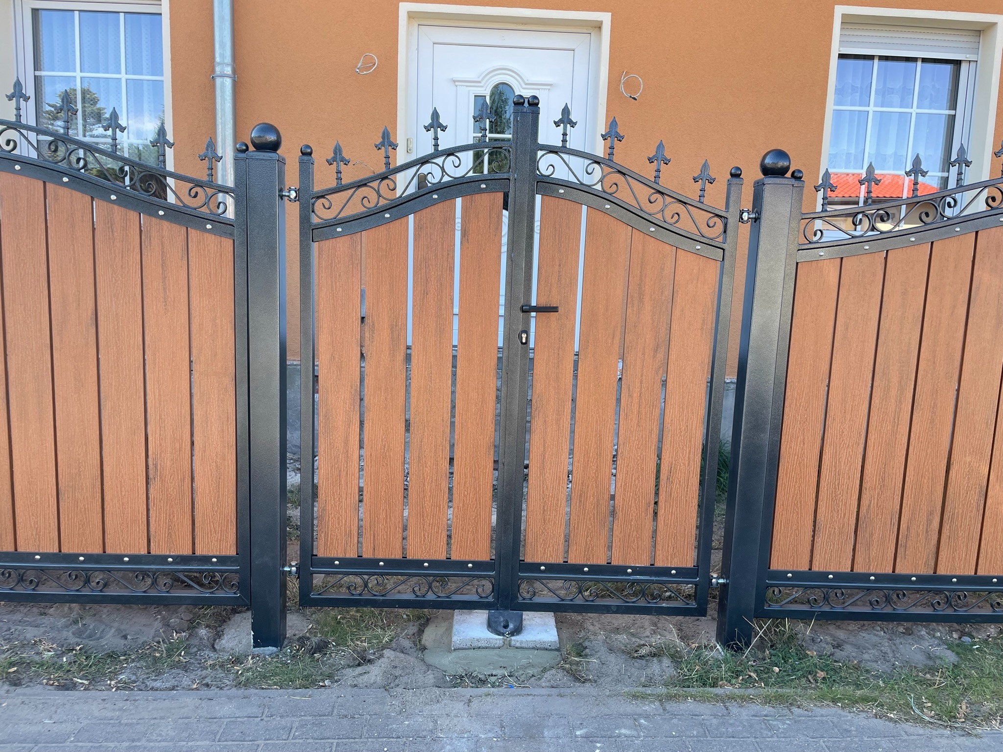 Dwuskrzydłowa brama wjazdowa z drewnopodobnym wypełnieniem i czarnymi, kutymi elementami dekoracyjnymi, widok z zewnątrz na tle pomarańczowej elewacji domu.