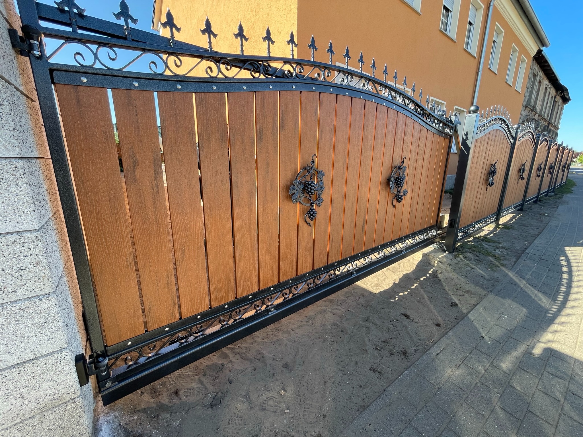 Ozdobna brama wjazdowa z drewnianymi panelami i metalowymi aplikacjami w kształcie winogron, widoczna z perspektywy ulicy.